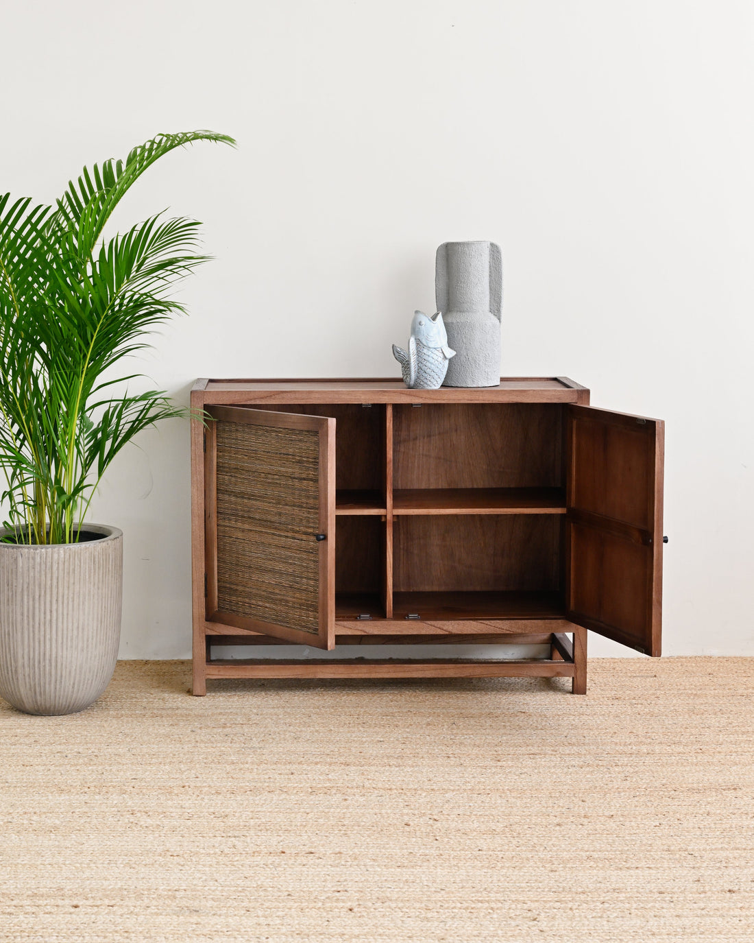A wooden sideboard cabinet with two open doors and open shelves, styled in a minimalist room with a potted palm plant and a small decorative vase on top.