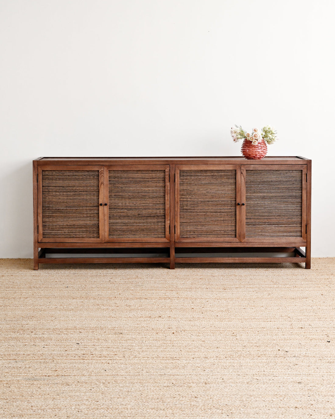 A mid-century modern wooden sideboard with woven rattan doors, set against a plain white wall on a light beige jute rug with a small terracotta vase of flowers on top.