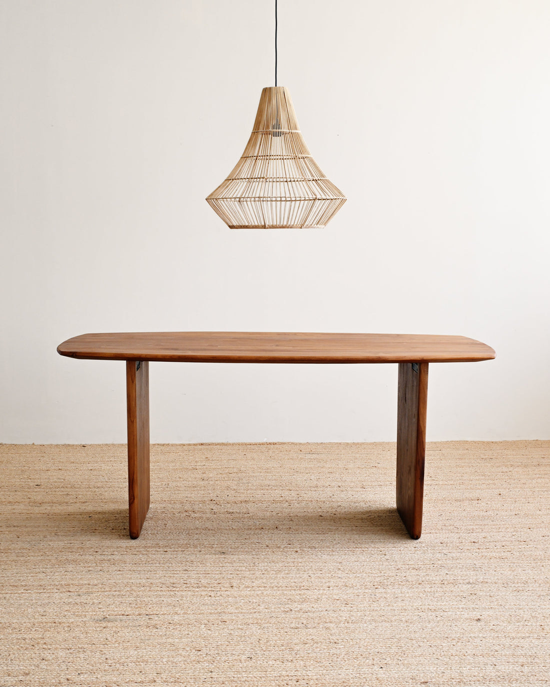 Wooden dining table with a hanging woven pendant light against a plain wall.