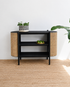 Black side table with wicker accents in a room with a white wall and wooden floor.