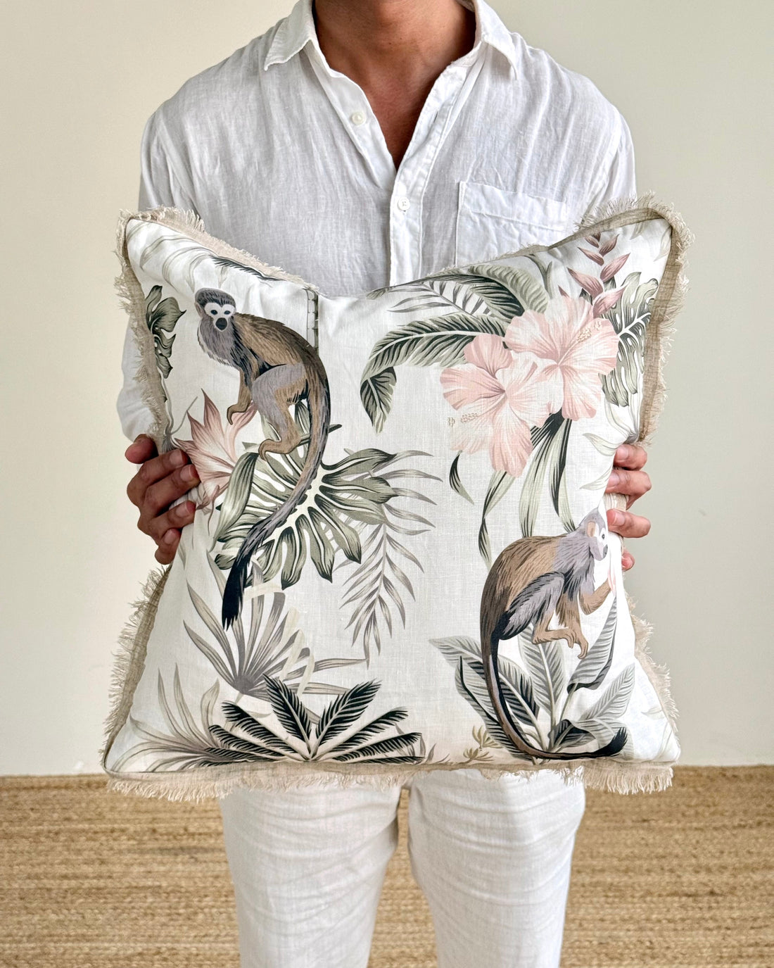 A person in a white linen shirt and white pants holds a square throw pillow featuring a monochrome and pastel jungle print with monkeys, palm leaves, and pink flowers.