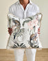 A person in a white linen shirt and white pants holds a square throw pillow featuring a monochrome and pastel jungle print with monkeys, palm leaves, and pink flowers.