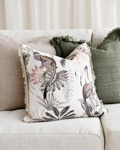 A square decorative throw pillow with a tropical bird and floral print in muted tones, styled on a light beige textured sofa alongside other neutral and green pillows.