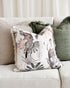 A square decorative throw pillow with a tropical bird and floral print in muted tones, styled on a light beige textured sofa alongside other neutral and green pillows.