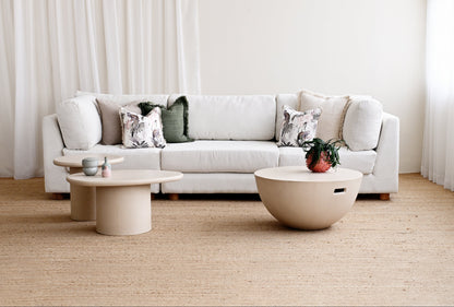 A modern light beige sectional sofa styled in a minimalist living room with neutral-toned coffee tables, patterned throw pillows, and sheer white curtains.