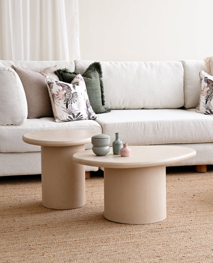 A styled living room scene featuring a white fabric sofa with decorative pillows, two round beige concrete coffee tables, and a textured beige rug on the floor.