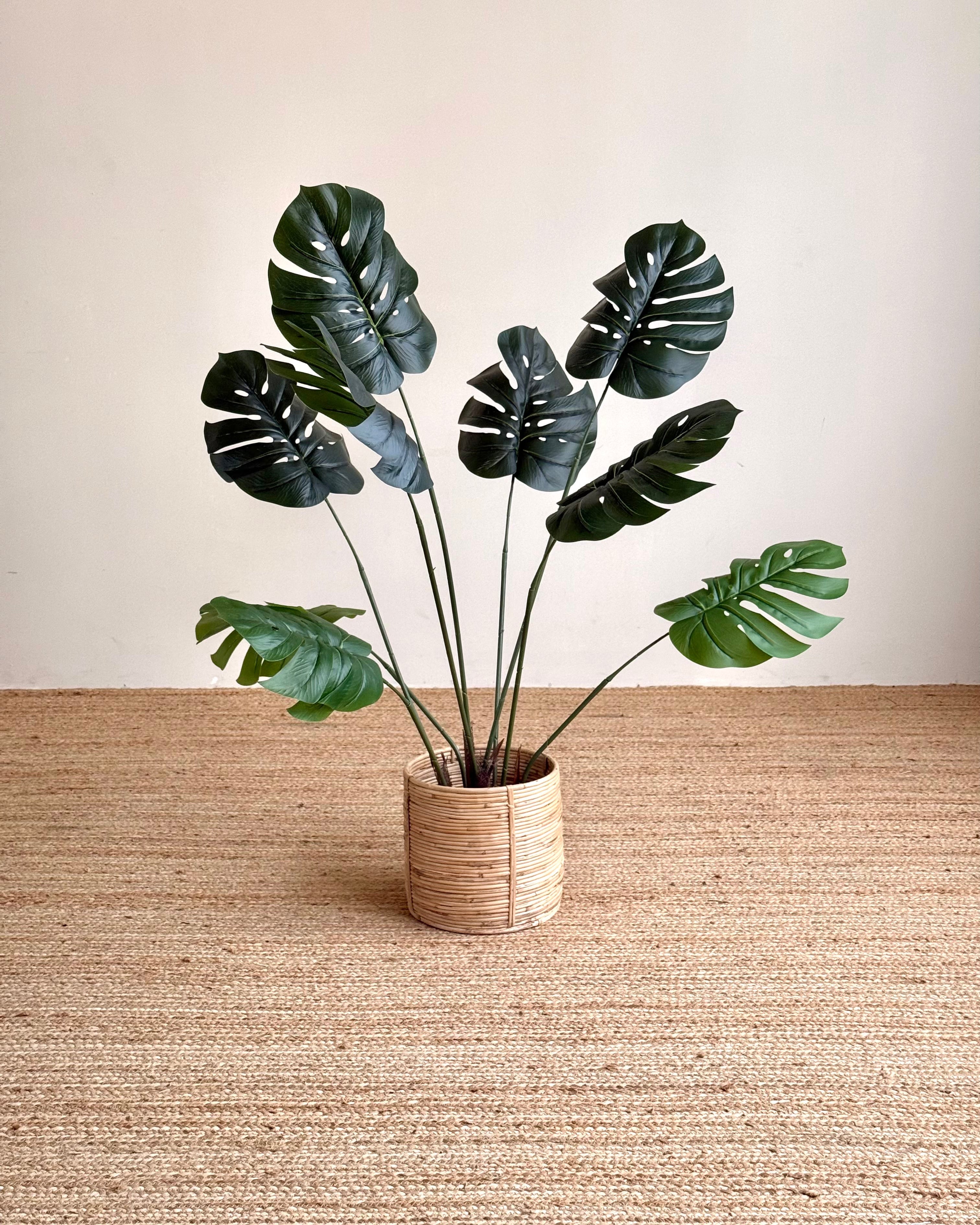 Artificial monstera plant with dark green leaves in a woven beige pot, set on a textured jute rug against a neutral beige wall.