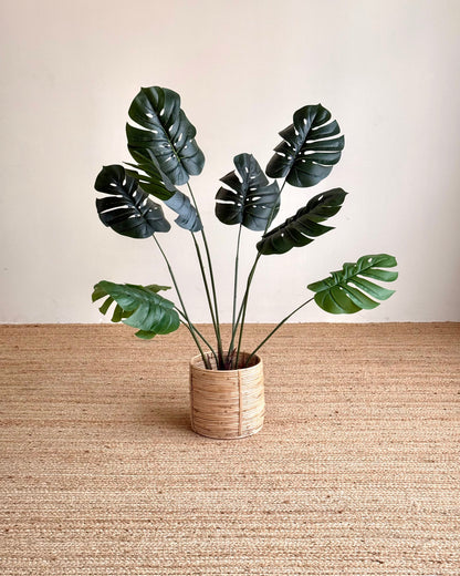 Artificial monstera plant with dark green leaves in a woven beige pot, set on a textured jute rug against a neutral beige wall.
