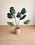 Artificial monstera plant with dark green leaves in a woven beige pot, set on a textured jute rug against a neutral beige wall.