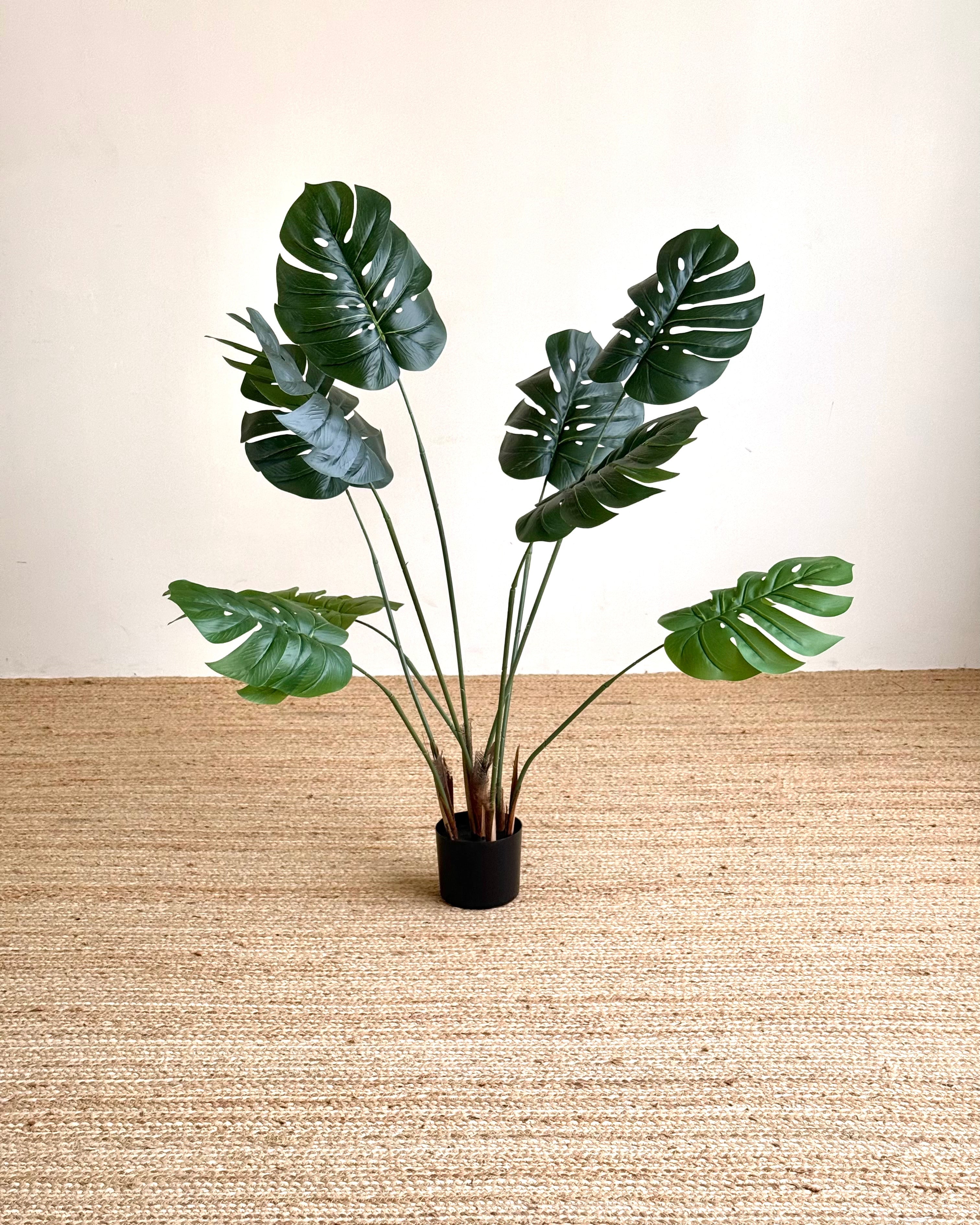 A potted artificial monstera plant with realistic green leaves and brown stems, set in a black pot on a beige woven rug against a cream wall.