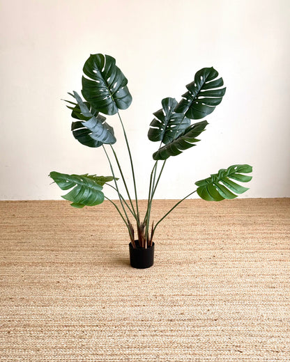 A potted artificial monstera plant with realistic green leaves and brown stems, set in a black pot on a beige woven rug against a cream wall.