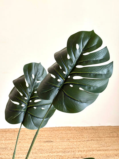 Two large artificial monstera deliciosa leaves with realistic fenestrations and glossy dark green surfaces, attached to long stems against a light cream wall and woven jute floor.