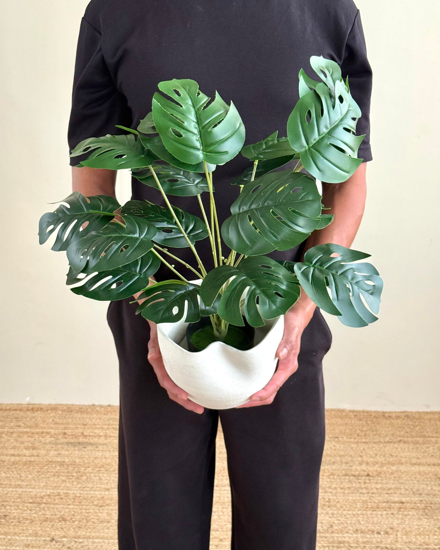 A person wearing a black t-shirt and black pants holds a potted artificial Monstera plant in a white wavy ceramic pot, standing against a beige wall and light brown woven floor.