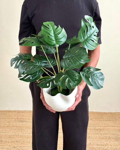 A person wearing a black t-shirt and black pants holds a potted artificial Monstera plant in a white wavy ceramic pot, standing against a beige wall and light brown woven floor.