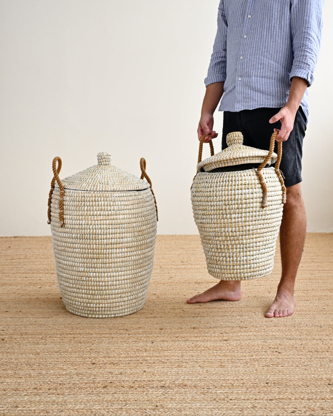 A man stands barefoot on a woven jute floor against a plain off-white wall, holding a large woven storage basket with a lid and rope handles. Another matching basket sits on the floor beside him.