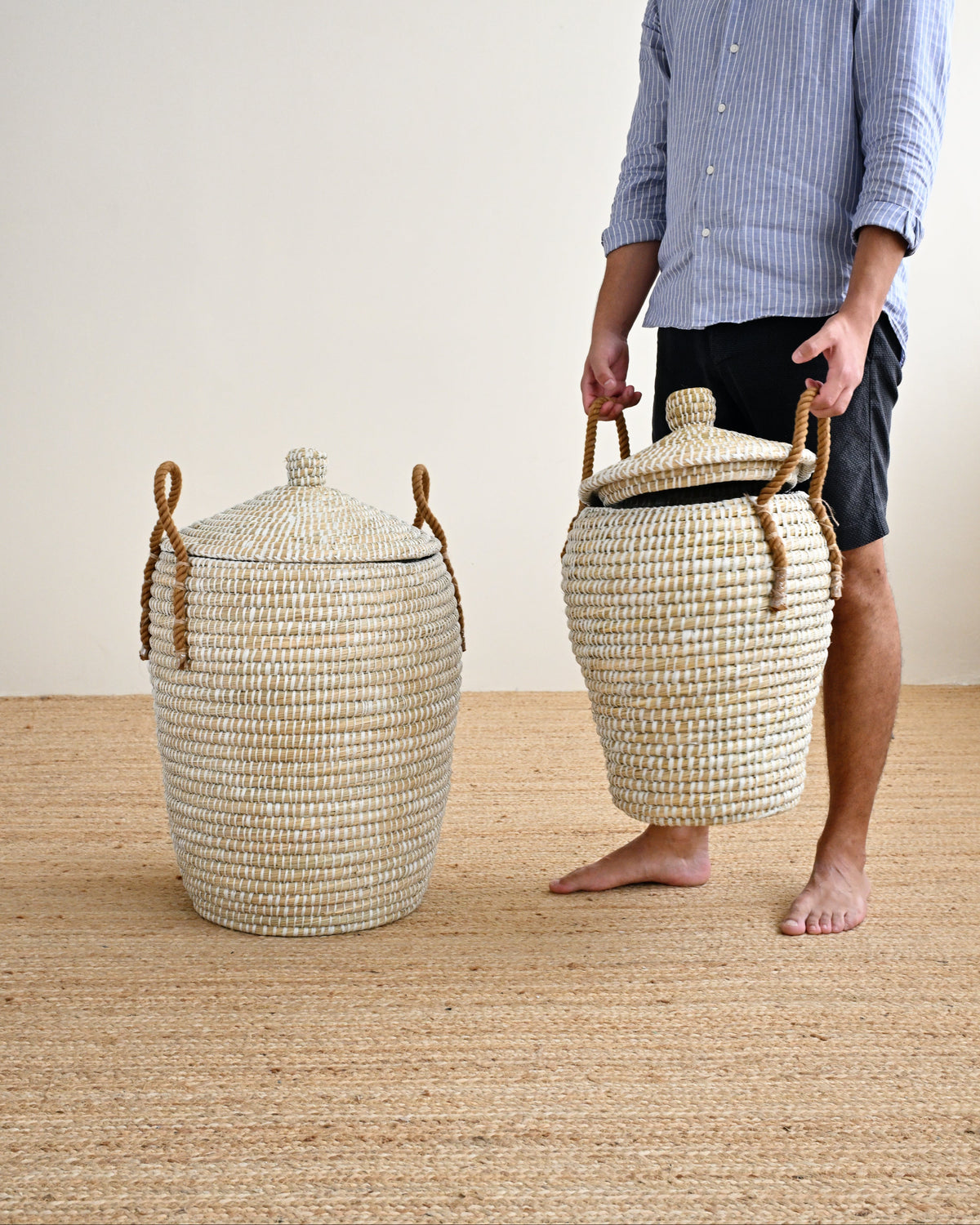 A man stands barefoot on a woven jute floor against a plain off-white wall, holding a large woven storage basket with a lid and rope handles. Another matching basket sits on the floor beside him.