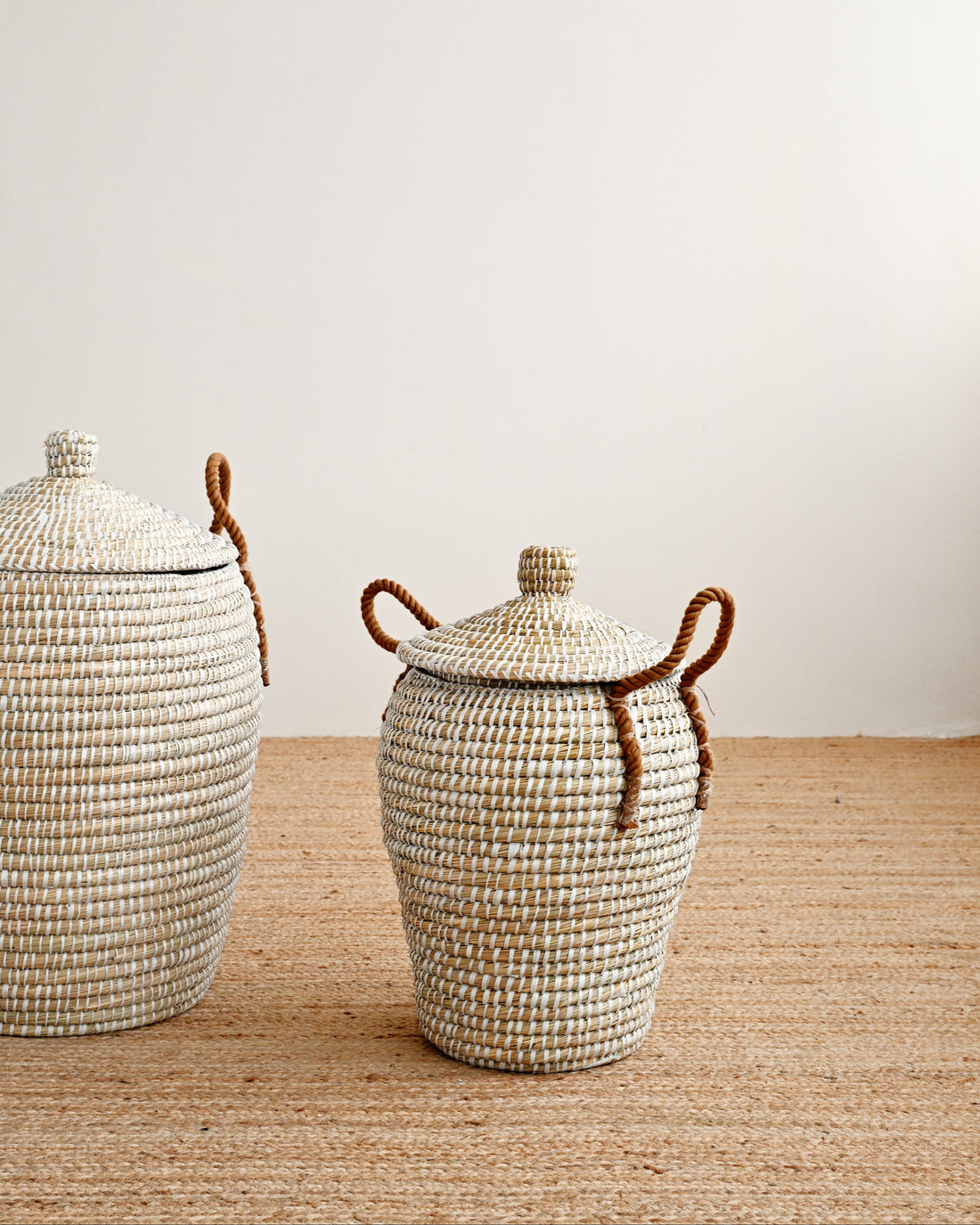 Two woven natural-fiber storage baskets with lids and brown rope handles, displayed on a woven rug against a plain off-white wall.