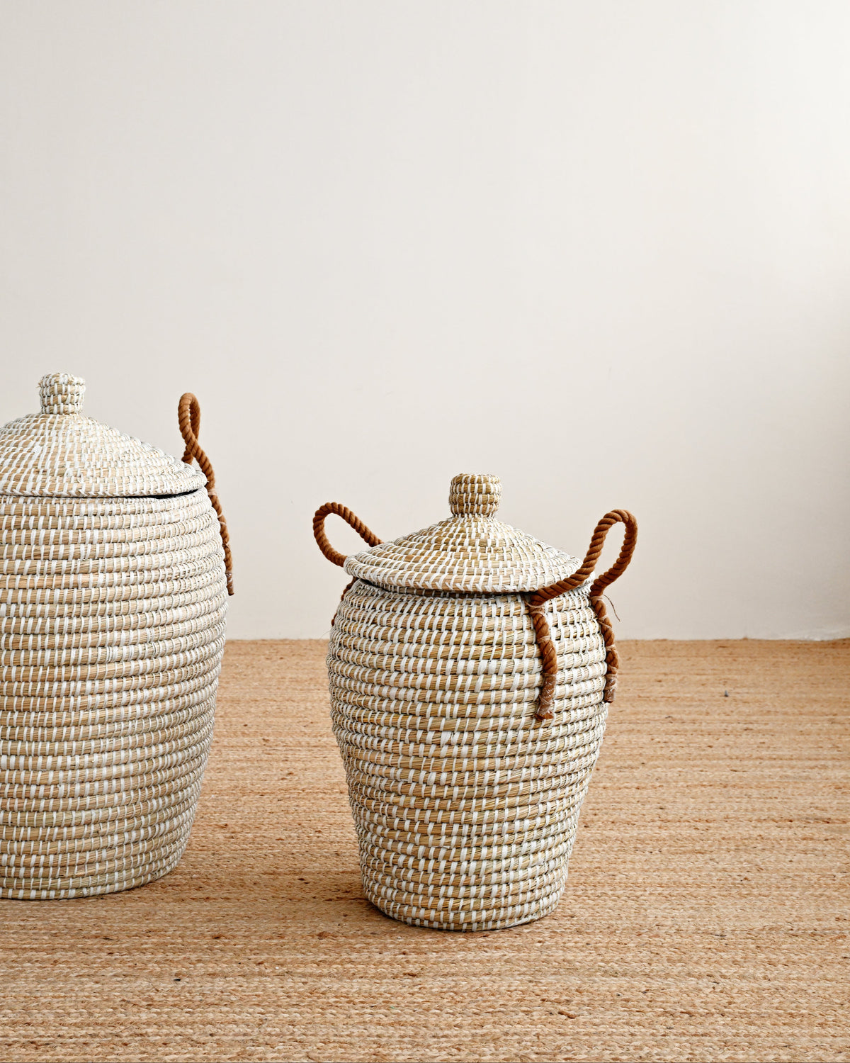Two woven natural-fiber storage baskets with lids and brown rope handles, displayed on a woven rug against a plain off-white wall.