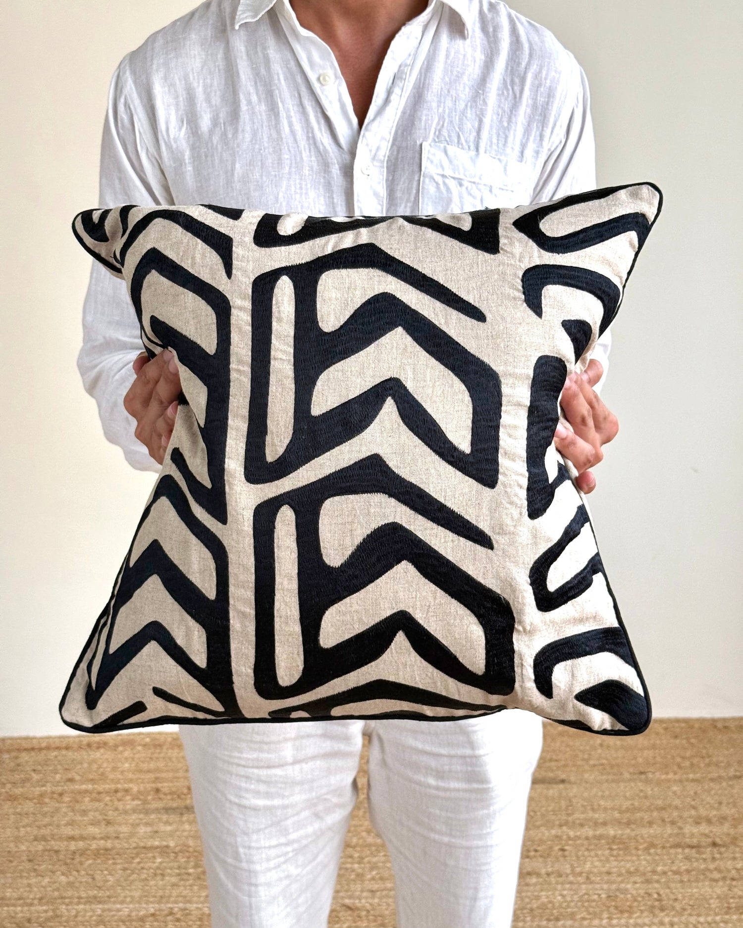 A person in a white linen shirt and white pants holds a square throw pillow with a bold black and cream geometric pattern.