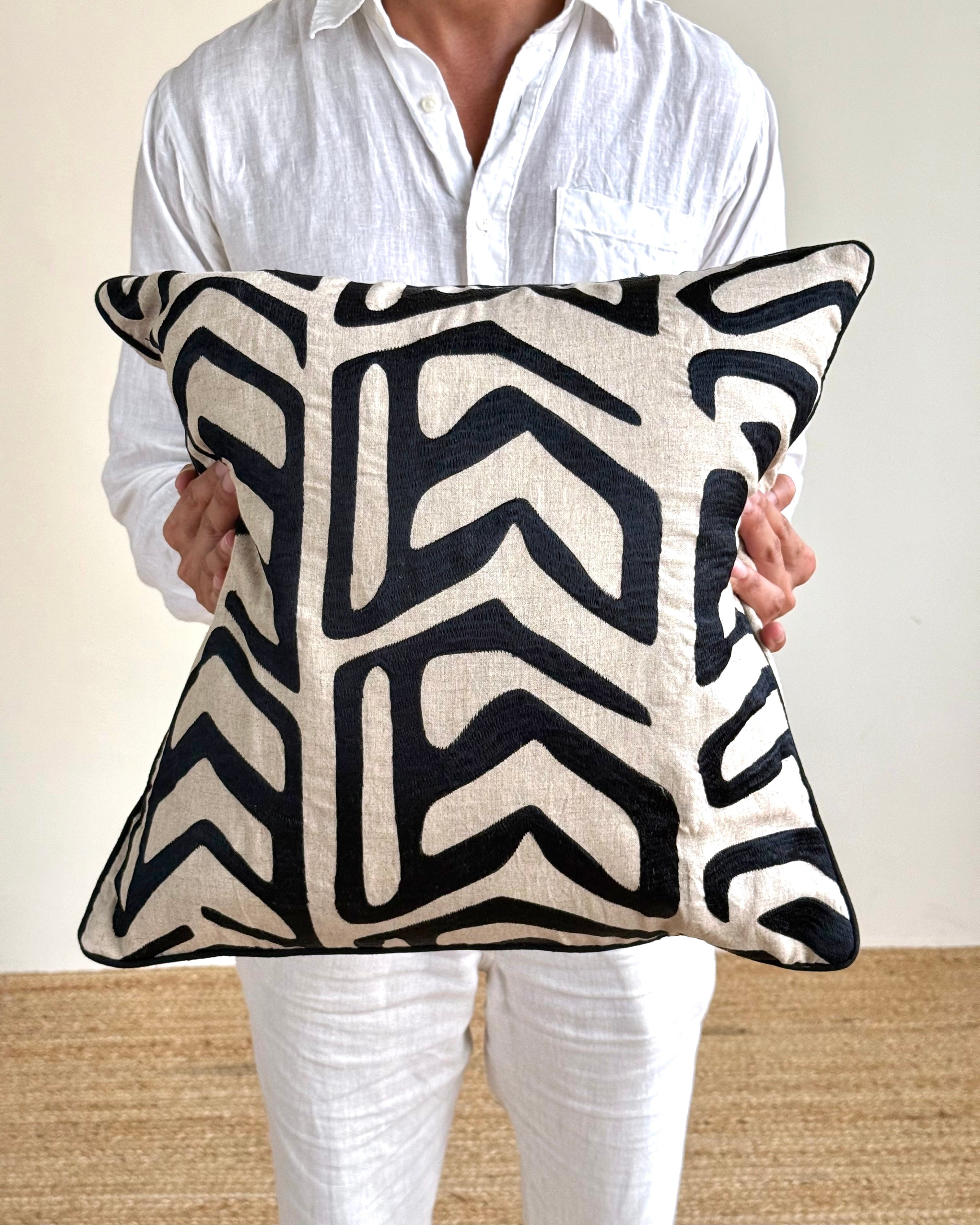 A person in a white linen shirt and white pants holds a square throw pillow with a bold black and cream geometric pattern.