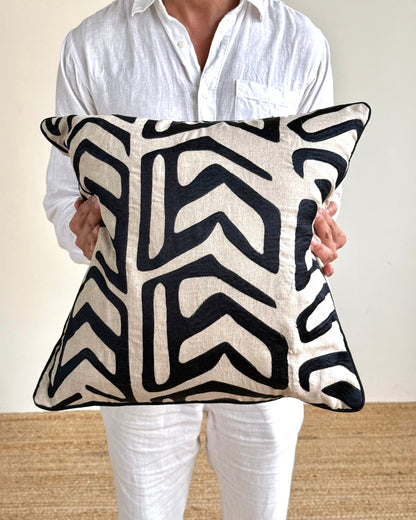 A person in a white linen shirt and white pants holds a square throw pillow with a bold black and cream geometric pattern.