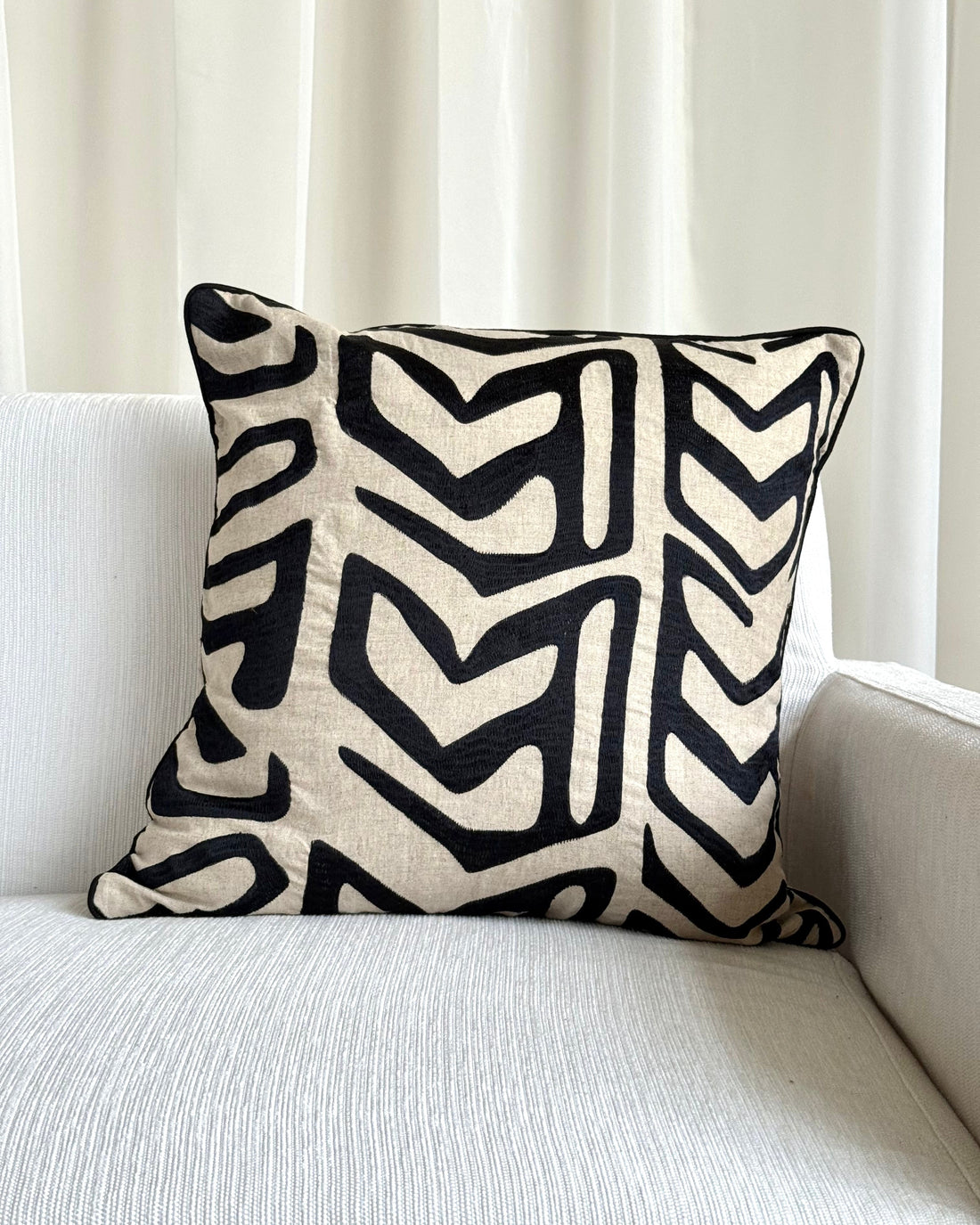 A square throw pillow with a bold black and cream geometric chevron pattern, resting on a light gray textured sofa in front of white curtains.