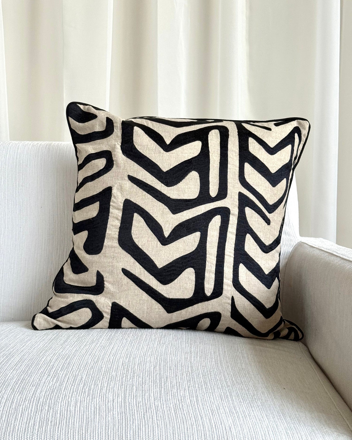 A square throw pillow with a bold black and cream geometric chevron pattern, resting on a light gray textured sofa in front of white curtains.