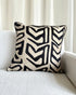 A square throw pillow with a bold black and cream geometric chevron pattern, resting on a light gray textured sofa in front of white curtains.