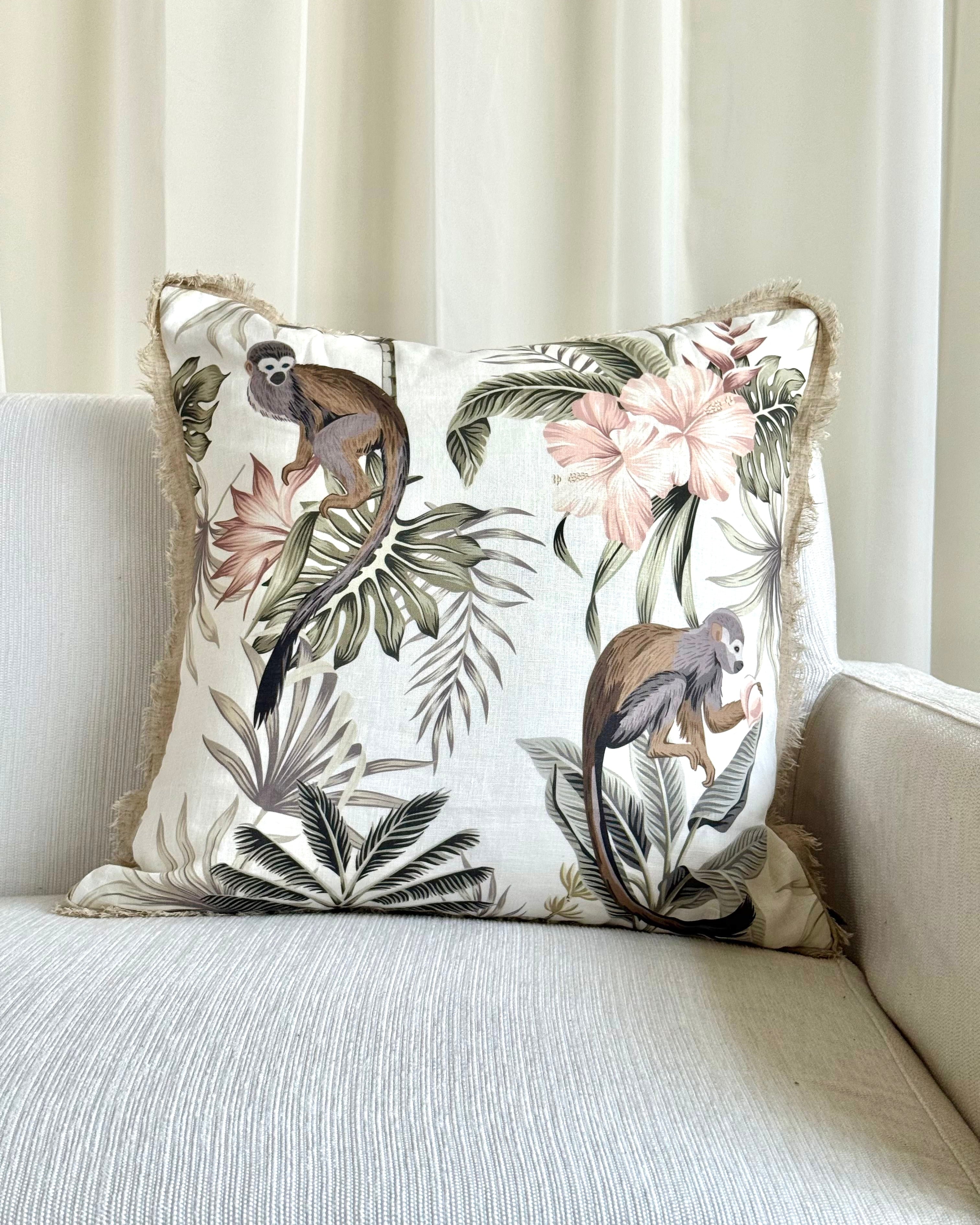 A square decorative throw pillow with a tropical jungle print featuring monkeys, palm leaves, and pink flowers, resting on a light beige textured sofa in front of cream curtains.
