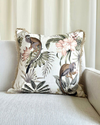 A square decorative throw pillow with a tropical jungle print featuring monkeys, palm leaves, and pink flowers, resting on a light beige textured sofa in front of cream curtains.