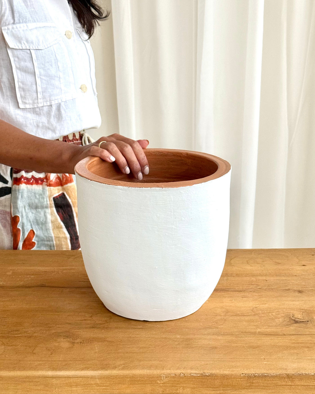 A person wearing a white sleeveless top and a patterned skirt stands behind a large white ceramic planter with a natural wood rim, placed on a wooden table in front of a white curtain.
