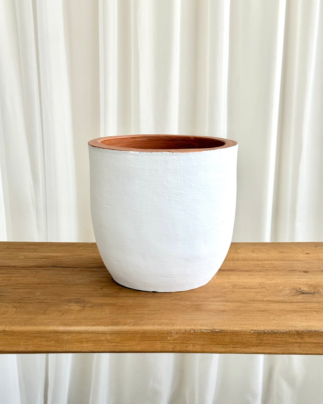 A white textured ceramic planter with a warm terracotta-colored interior, displayed on a rustic wooden table against a white curtain background.
