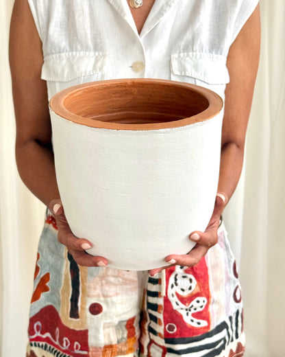 A person holding a large cylindrical white planter pot with a terracotta-colored interior, wearing a white sleeveless top and patterned shorts.