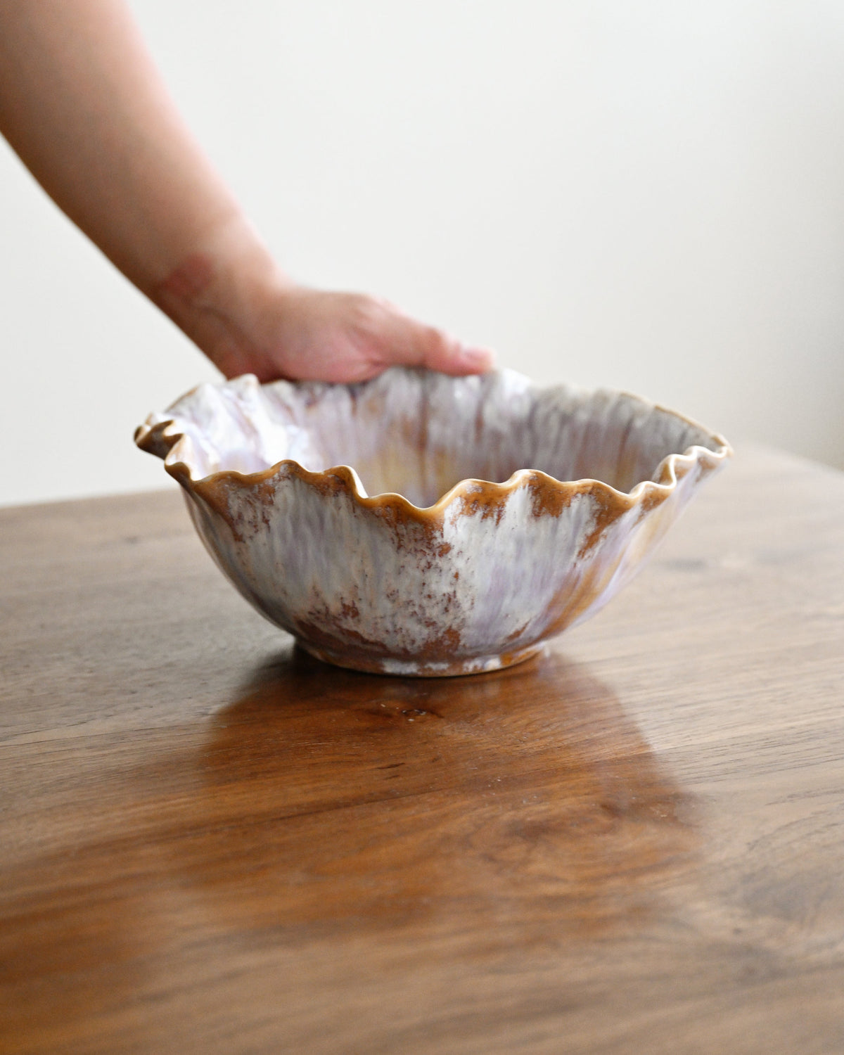 A hand holding a scalloped ceramic bowl with a rustic brown and cream glaze, resting on a wooden table against a plain light background.