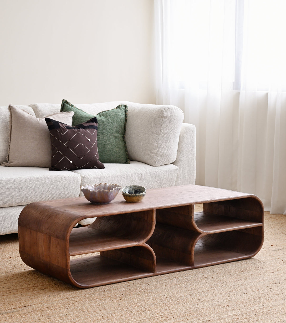 A curved wooden coffee table with open shelves and two small bowls on top, placed in a bright living room in front of a white sofa with decorative pillows.