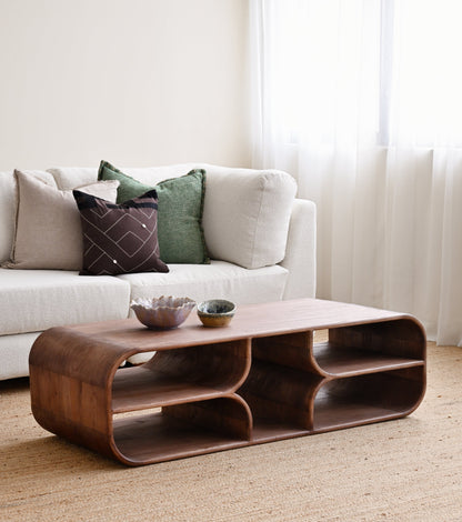 A curved wooden coffee table with open shelves and two small bowls on top, placed in a bright living room in front of a white sofa with decorative pillows.