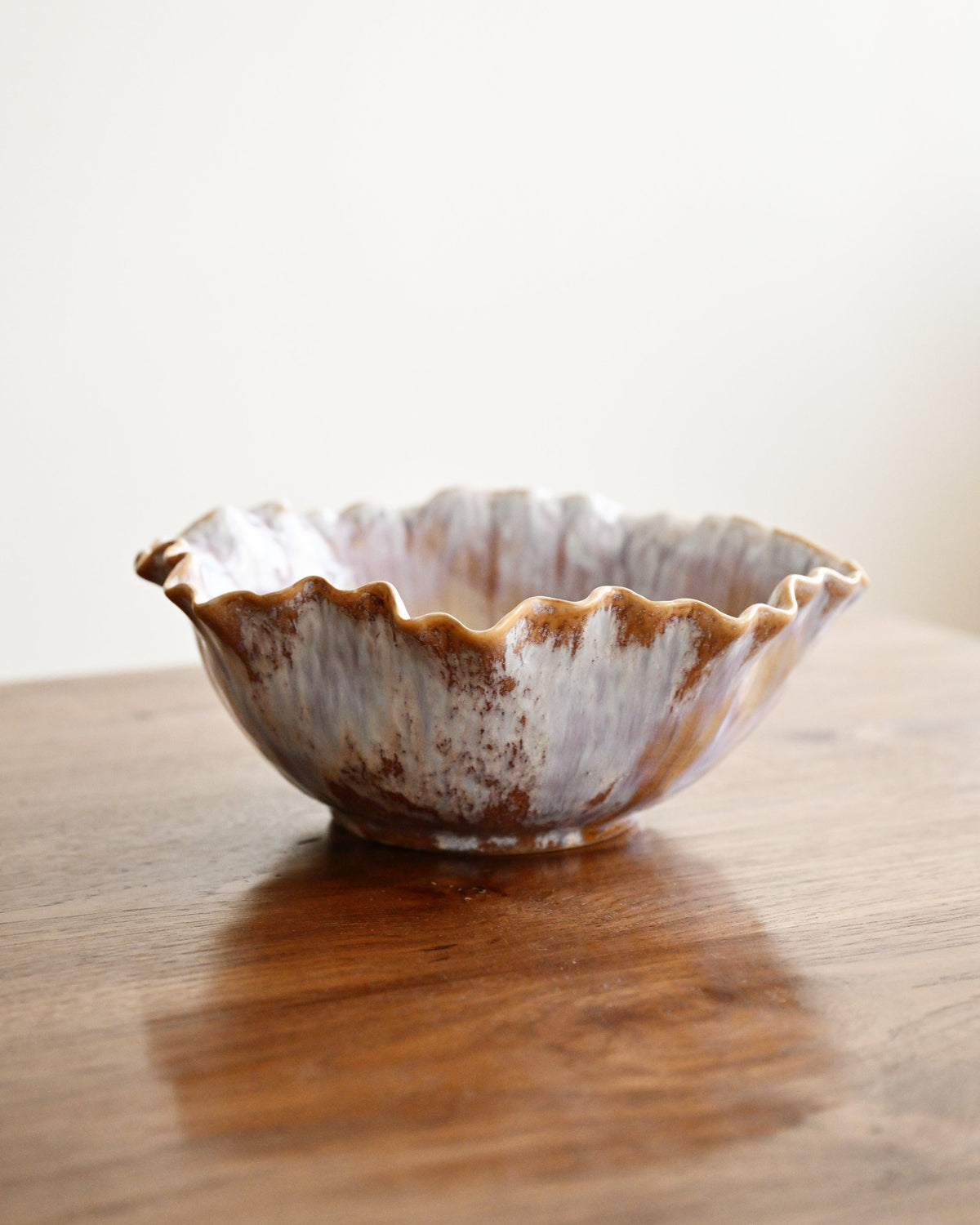 A handmade ceramic bowl with a scalloped rim, featuring a rustic glaze of brown and off-white speckled patterns, sitting on a wooden table against a plain light background.