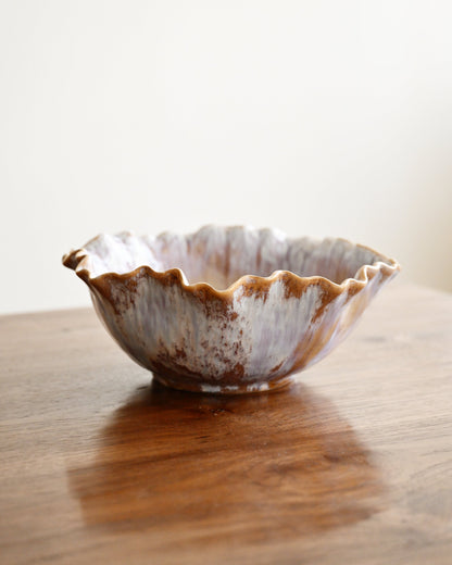 A handmade ceramic bowl with a scalloped rim, featuring a rustic glaze of brown and off-white speckled patterns, sitting on a wooden table against a plain light background.