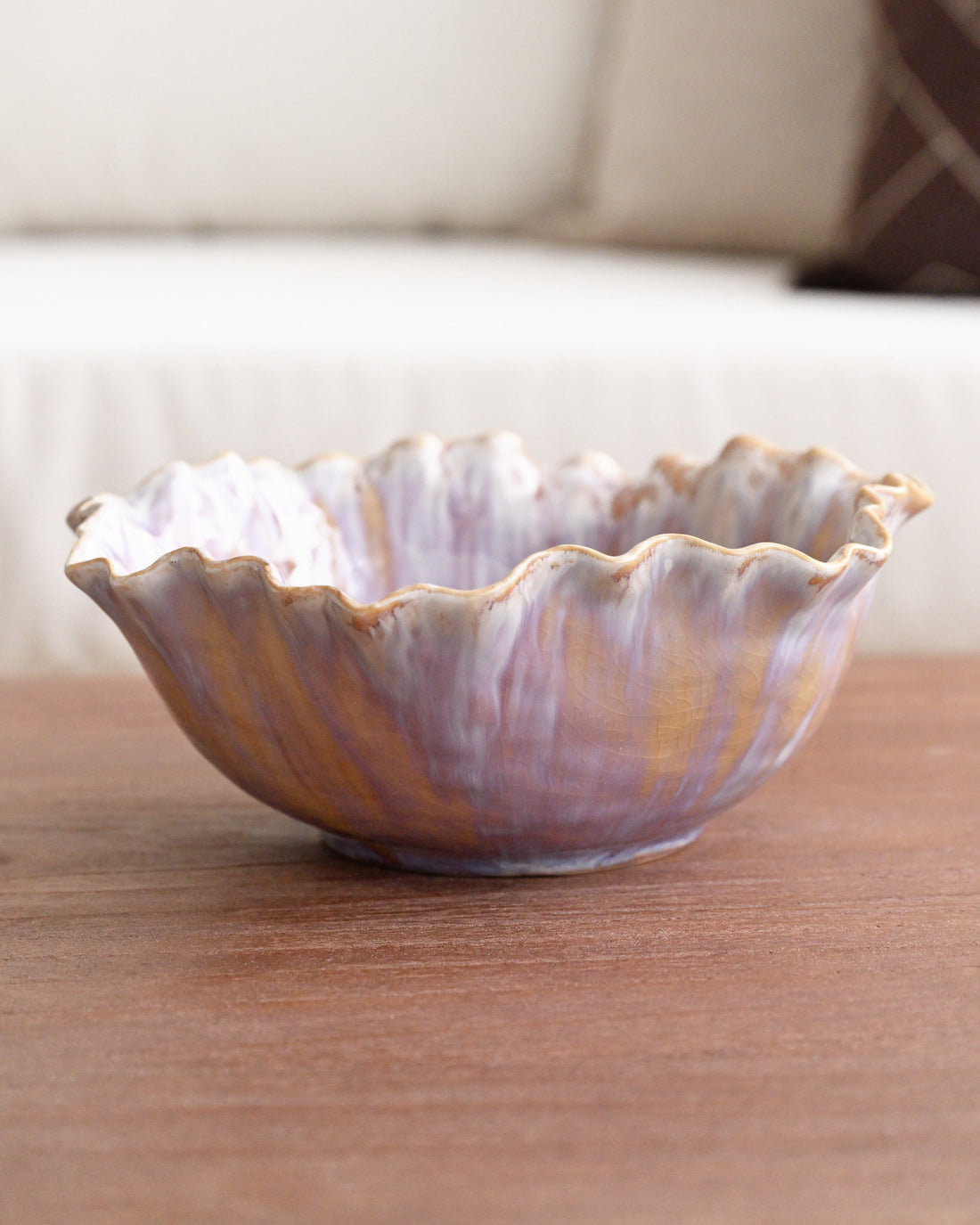 A scalloped ceramic bowl with a marbled purple, gold, and white glaze, resting on a wooden table with a blurred sofa in the background.