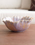 A scalloped ceramic bowl with a marbled purple, gold, and white glaze, resting on a wooden table with a blurred sofa in the background.