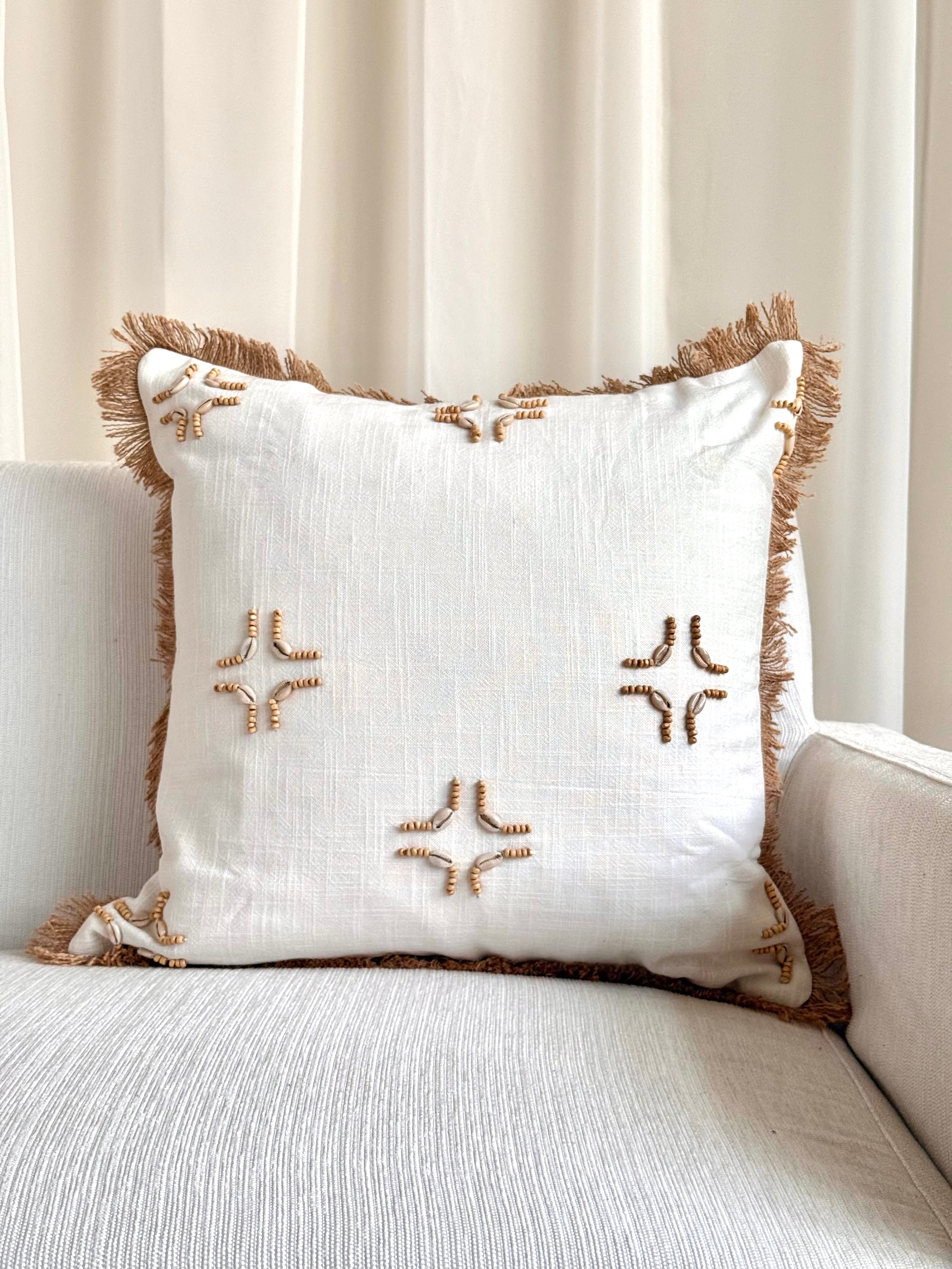 A square off-white throw pillow with brown fringe trim and embroidered starburst motifs, resting on a light beige sofa in front of cream curtains.