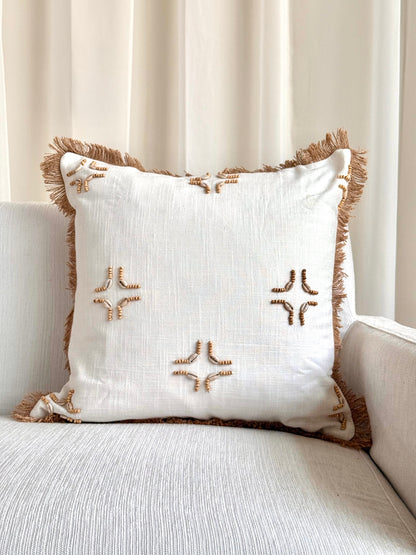 A square off-white throw pillow with brown fringe trim and embroidered starburst motifs, resting on a light beige sofa in front of cream curtains.