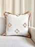 A square off-white throw pillow with brown fringe trim and embroidered starburst motifs, resting on a light beige sofa in front of cream curtains.