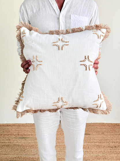A person in a white linen shirt and white pants holds a square white throw pillow with fringed edges and tan embroidered geometric patterns.