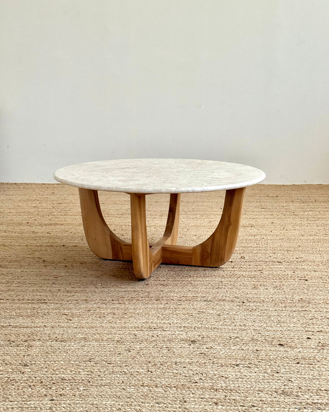 A round coffee table with a white marble top and a curved natural wood base, set on a woven jute rug against a plain off-white wall.