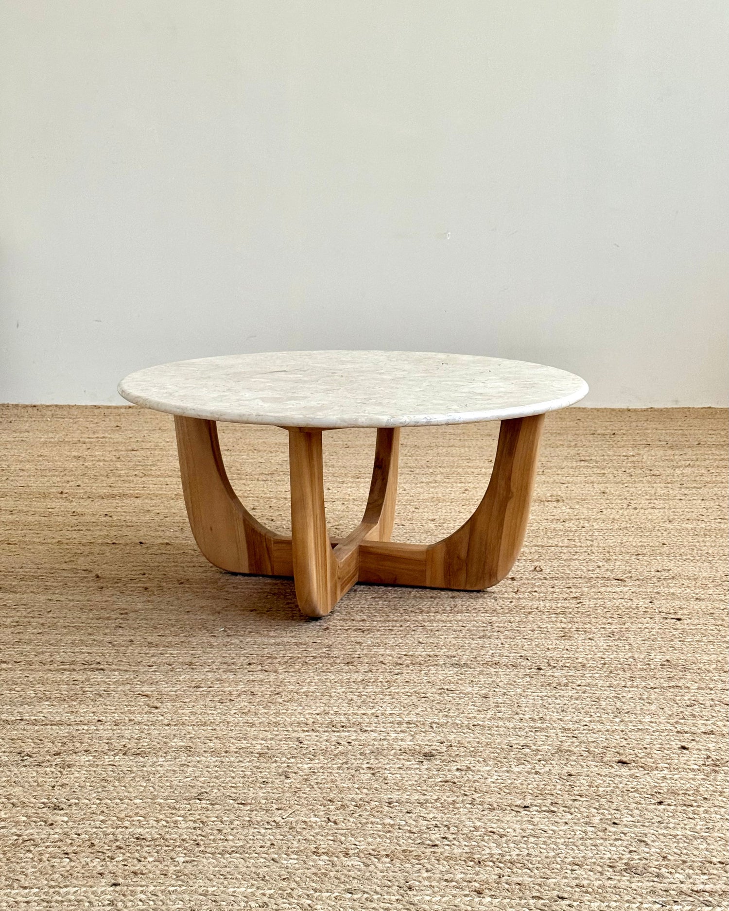 A round coffee table with a white marble top and a curved natural wood base, set on a woven jute rug against a plain off-white wall.