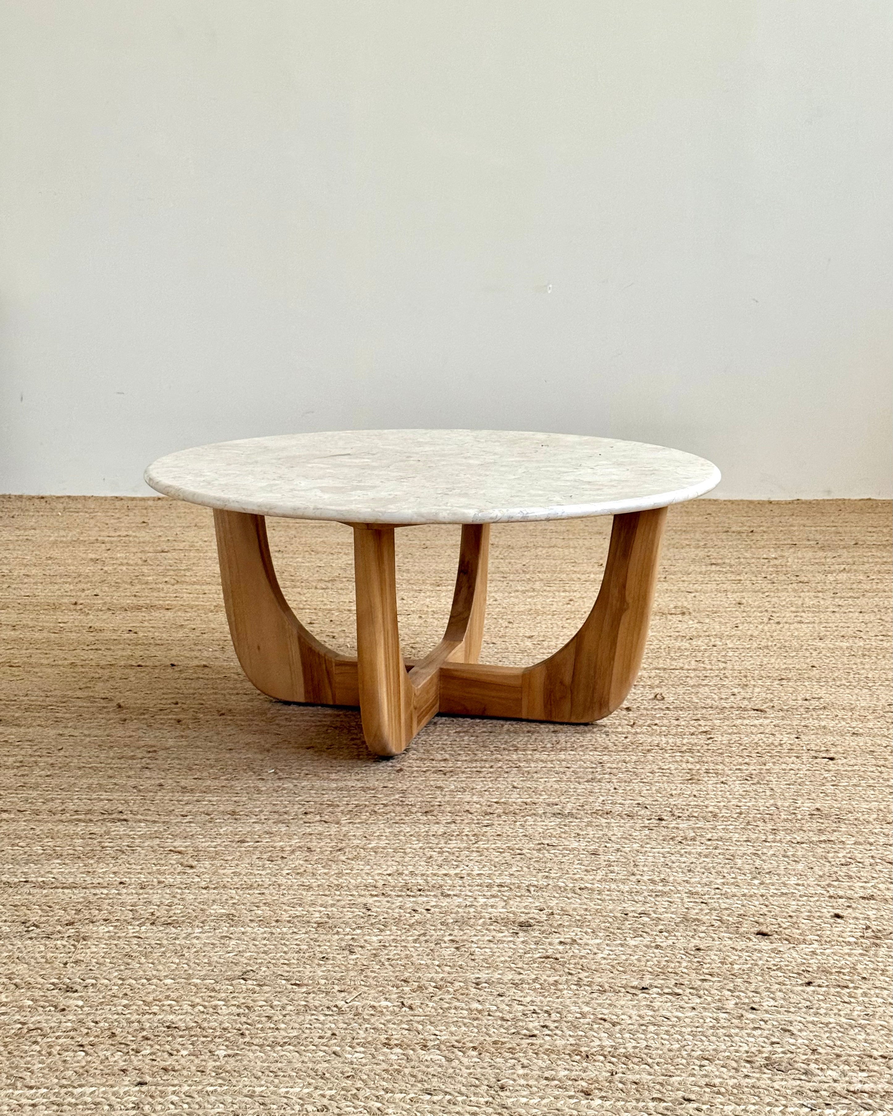A round coffee table with a white marble top and a curved natural wood base, set on a woven jute rug against a plain off-white wall.