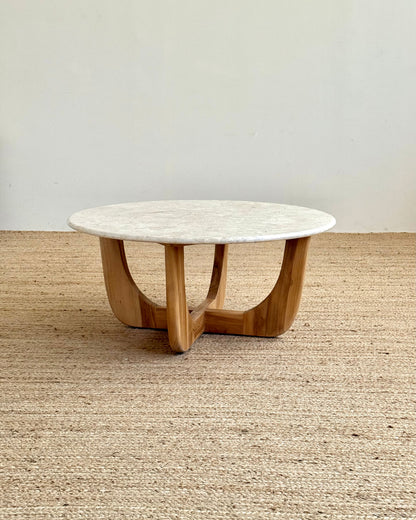 A round coffee table with a white marble top and a curved natural wood base, set on a woven jute rug against a plain off-white wall.