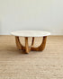 A round coffee table with a white marble top and a curved natural wood base, set on a woven jute rug against a plain off-white wall.