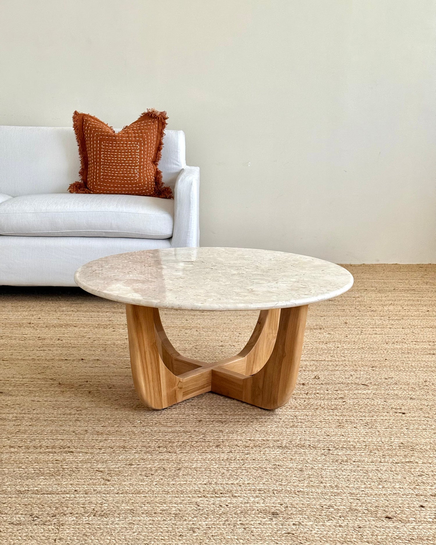 A round coffee table with a light beige stone top and a natural wood base, styled in a living room setting on a woven jute rug with a white sofa in the background.
